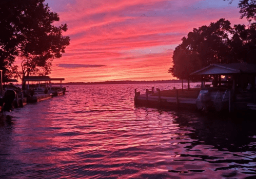 A vibrant sunset over a lake, with pink and orange hues reflecting on the water and silhouettes of trees and boats.