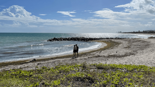 A couple stands on a sandy beach, gazing at the ocean under a partly cloudy sky.