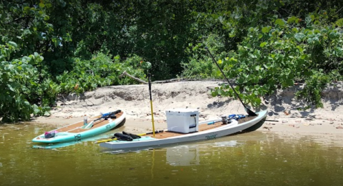 Two kayaks are anchored by a sandy bank, surrounded by lush greenery, with fishing gear and a cooler on board.
