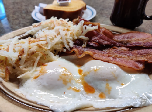 A plate of breakfast featuring two fried eggs, crispy bacon, hash browns, and a slice of toast in the background.