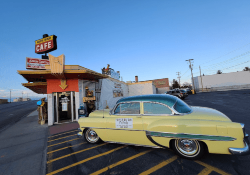 A vintage yellow car parked outside a retro diner with a sign reading "North Way Cafe" against a clear blue sky.
