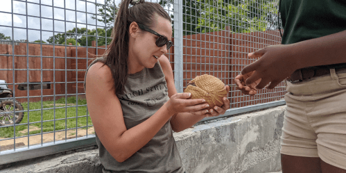 A woman in sunglasses examines a textured object while another person hands it to her, with a fenced area in the background.