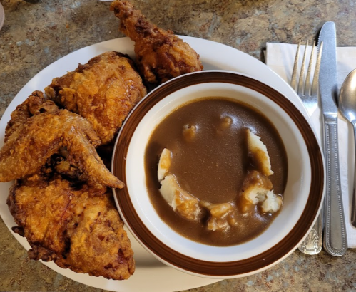 A plate of crispy fried chicken with a bowl of brown gravy and chunks of mashed potatoes on a textured surface.