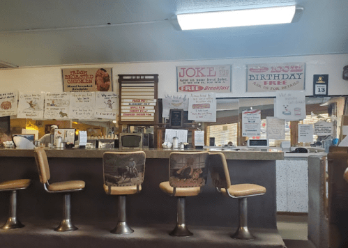 A diner interior with a counter, stools, and various humorous signs on the walls promoting food and events.