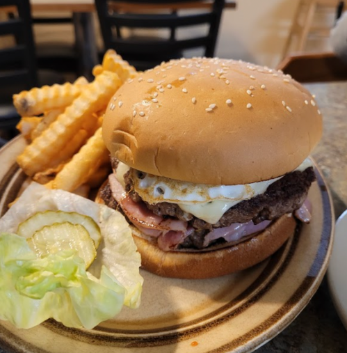 A sesame seed bun burger with ham, a fried egg, lettuce, and pickles, served with crinkle-cut fries on a plate.