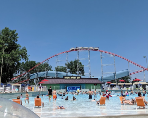 A water park scene with a wave pool, sunbathers, and a roller coaster in the background under a clear blue sky.