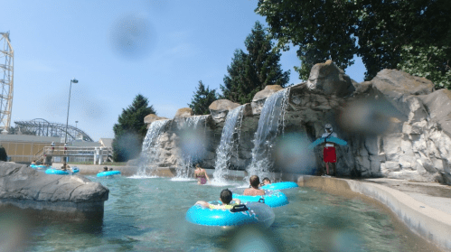 People float on inner tubes in a pool with waterfalls and rocks under a clear blue sky.