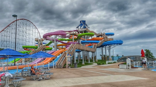 A colorful water park with multiple slides, a roller coaster in the background, and people relaxing under umbrellas.