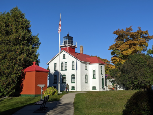 A white lighthouse with a red roof, surrounded by greenery and trees, under a clear blue sky.