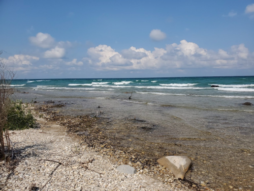 A serene beach scene with gentle waves, rocky shore, and a clear blue sky dotted with clouds.