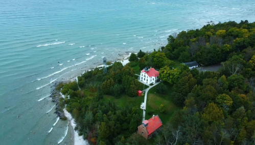 Aerial view of a coastal landscape featuring a lighthouse, red-roofed buildings, and waves lapping at the shore.