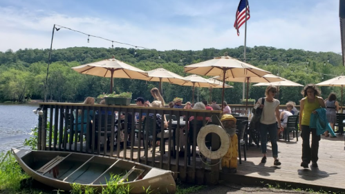 A riverside deck with umbrellas, people dining, and a small boat nearby, set against a green hillside and blue sky.