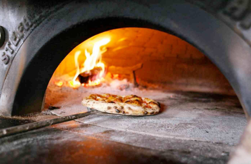 A freshly baked pizza being pulled from a wood-fired oven, with flames visible in the background.