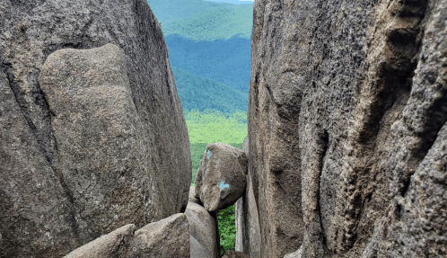 A narrow gap between large rocks, with lush green mountains visible in the background under a clear sky.