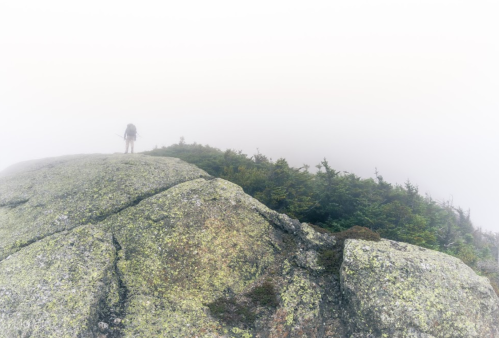 A lone hiker stands on a rocky, fog-covered mountain peak surrounded by trees.
