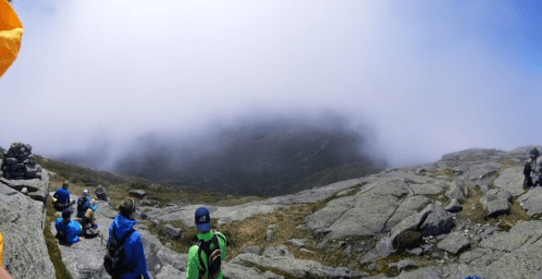 A group of hikers rests on rocky terrain, surrounded by fog and mountains in the background.