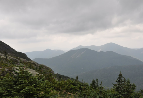 A panoramic view of misty mountains under a cloudy sky, with green foliage in the foreground.