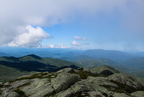 A panoramic view of mountains under a partly cloudy sky, with rocky terrain in the foreground and distant peaks.