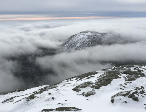 A snowy mountain landscape with clouds rolling over the peaks under a gray sky.