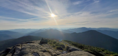 A panoramic view of mountains under a bright sun, with a clear sky and soft clouds in the background.