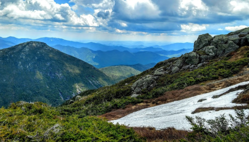 A scenic mountain landscape with rocky terrain, green shrubs, and patches of snow under a cloudy blue sky.