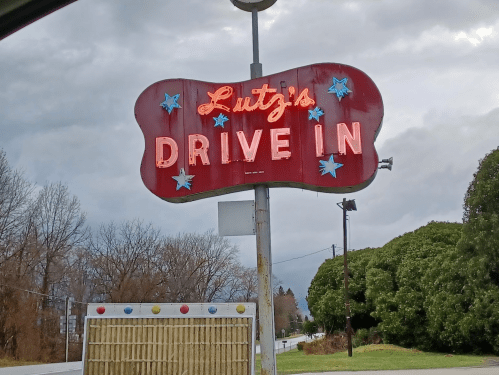 A vintage neon sign for "Lutz's Drive In" with stars, set against a cloudy sky and trees in the background.