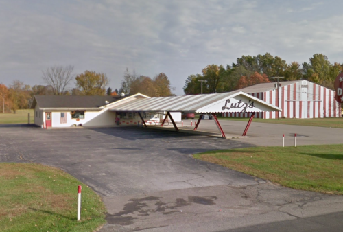 A small building with a large awning labeled "Lu's," set in a grassy area with trees and a striped building in the background.