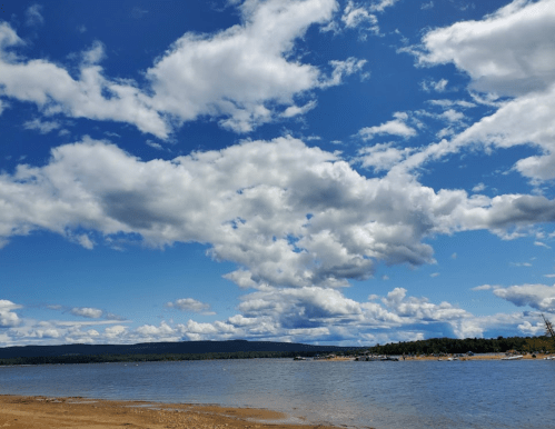 A serene lakeside view with a sandy shore, blue sky, and fluffy white clouds reflecting on the water.