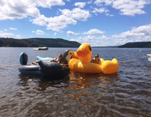 Two people relax on inflatable floats in a lake, surrounded by trees and a blue sky with fluffy clouds.
