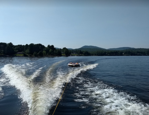 A boat pulls a tube across a calm lake, with mountains and trees in the background under a clear blue sky.