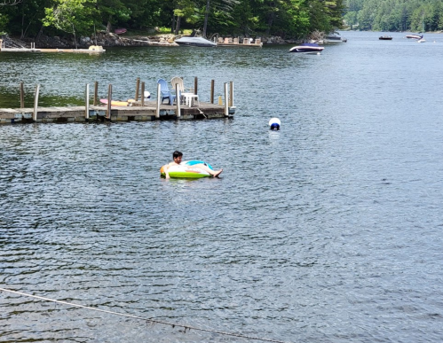 A person relaxes on a float in a calm lake, with a dock and boats visible in the background.
