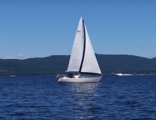 A white sailboat glides across a calm blue lake with mountains in the background under a clear blue sky.
