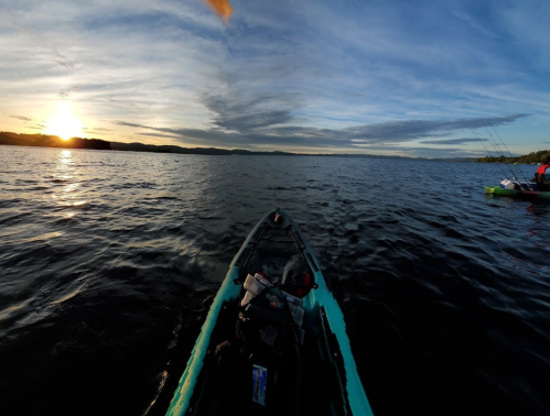 A kayak on calm water at sunset, with a distant silhouette of another kayak and a colorful sky.