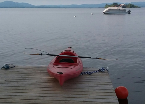 A red kayak on a wooden dock, with a boat in the background and calm water reflecting the sky.
