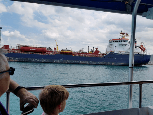 A child watches a large cargo ship from a boat, with cloudy skies and water in the background.