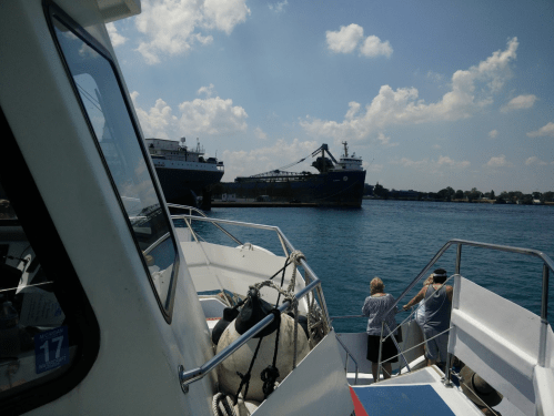 A view from a boat showing a large cargo ship and two people on the deck, with blue water and clouds in the background.