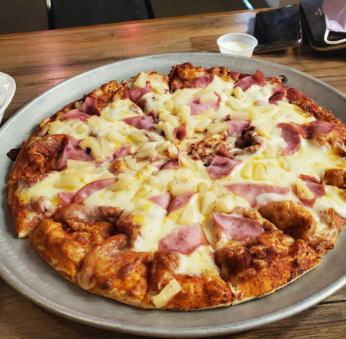 A large pizza topped with ham, pineapple, and melted cheese on a metal tray, set on a wooden table.