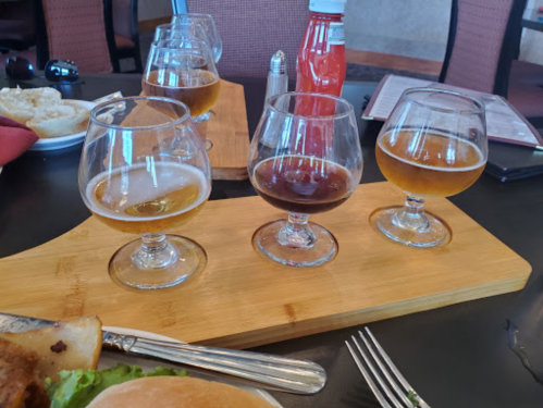 Three glasses of beer on a wooden board, with a plate of food and condiments in the background.