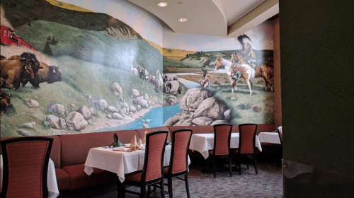 A restaurant interior featuring a mural of bison and horse riders by a river, with tables set for dining.
