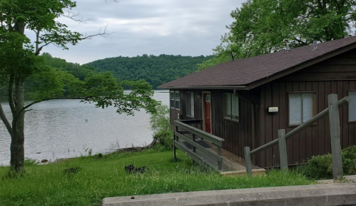 A brown cabin by a calm lake, surrounded by green trees and hills under a cloudy sky.