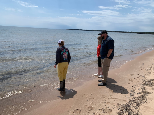 Three people stand on a sandy beach, looking out at the water under a partly cloudy sky.