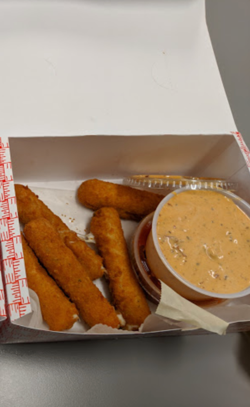 A takeout box containing several golden-brown fried sticks and a small cup of dipping sauce.