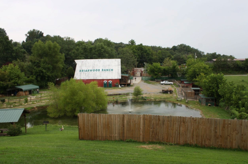 A scenic view of Briarwood Ranch featuring a pond, greenery, and rustic buildings surrounded by a wooden fence.