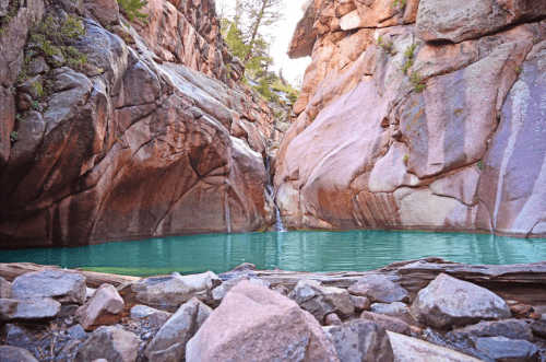 A serene canyon with towering rock walls and a turquoise pool of water at the base, surrounded by scattered stones.