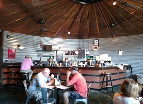 A cozy bar with a circular wooden ceiling, patrons seated at tables, and a bartender serving drinks.