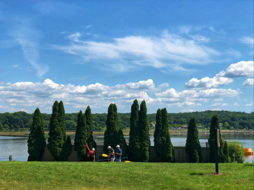 Two people prepare kayaks by a lake, with tall trees and a blue sky filled with clouds in the background.