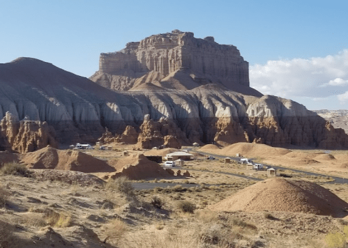 A desert landscape featuring a large rock formation, layered cliffs, and scattered small structures in the foreground.