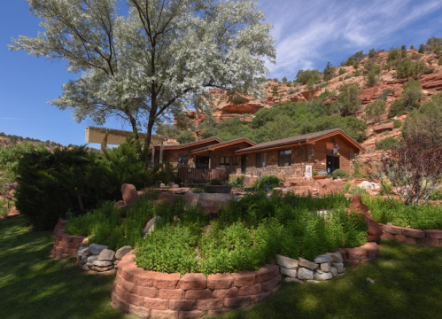 A rustic house surrounded by lush greenery and rocky hills under a clear blue sky.