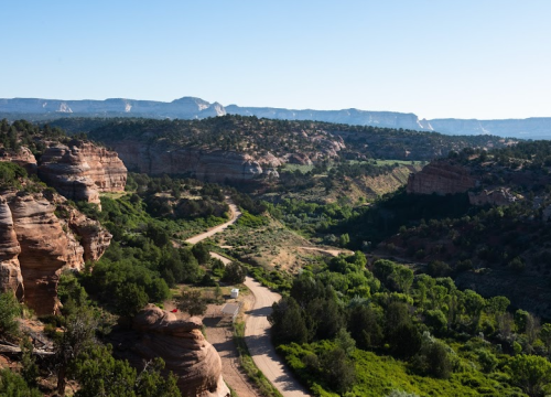 A winding dirt road through a lush green valley surrounded by rocky cliffs and distant mountains under a clear blue sky.