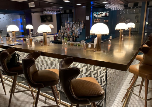 A stylish bar with a golden countertop, illuminated lamps, and high stools, featuring a well-stocked liquor display.